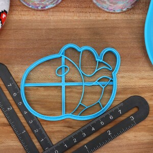May include: A blue plastic cookie cutter shaped like a cartoon character with a large head and a small body. The cutter has a grid pattern on the head and a curved line on the body.
