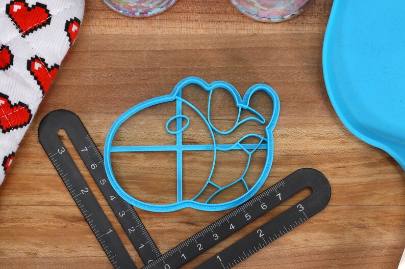 May include: A blue plastic cookie cutter shaped like a cartoon character with a large head and a small body. The cutter has a grid pattern on the head and a curved line on the body.
