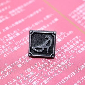 May include: A silver-toned metal pin with a square frame and a raised image of a high-heeled shoe inside. The pin has a slightly distressed finish.