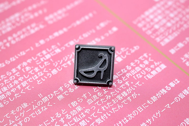 May include: A silver-toned metal pin with a square frame and a raised image of a high-heeled shoe inside. The pin has a slightly distressed finish.