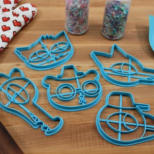 May include: A set of five blue plastic cookie cutters shaped like cartoon characters. The characters include a cat, a wolf, a bear wearing a hat and glasses, a character with a hat and a character with a hat and a long nose.