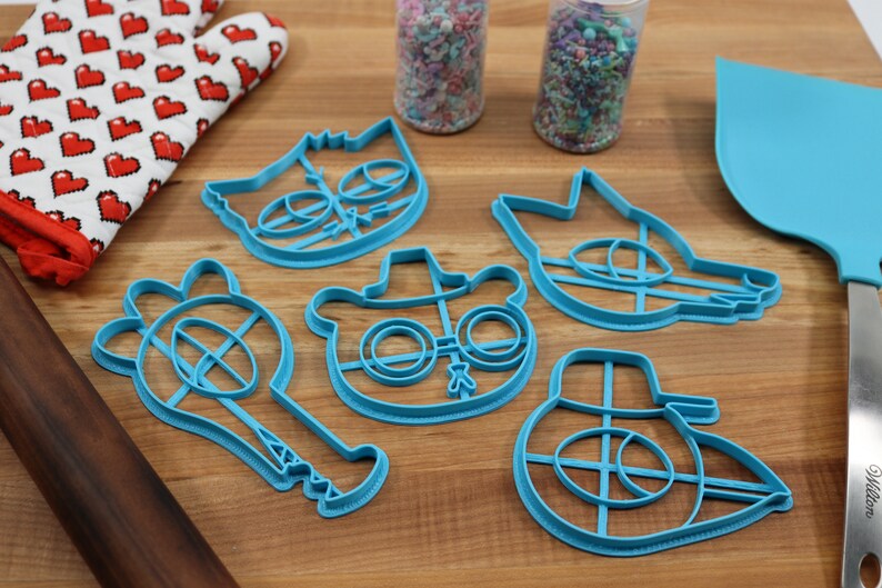 May include: A set of five blue plastic cookie cutters shaped like cartoon characters. The characters include a cat, a wolf, a bear wearing a hat and glasses, a character with a hat and a character with a hat and a long nose.