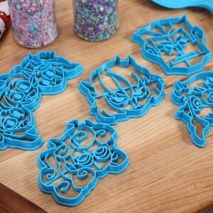 May include: Five blue plastic cookie cutters shaped like cartoon characters. The cutters are on a wooden surface.