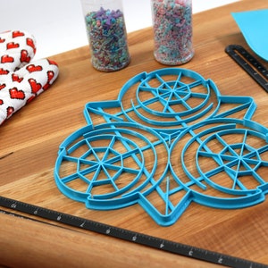 May include: A blue plastic cookie cutter shaped like three overlapping circles with a web-like design. The cutter is on a wooden surface with a rolling pin, a spatula, and two jars of sprinkles.