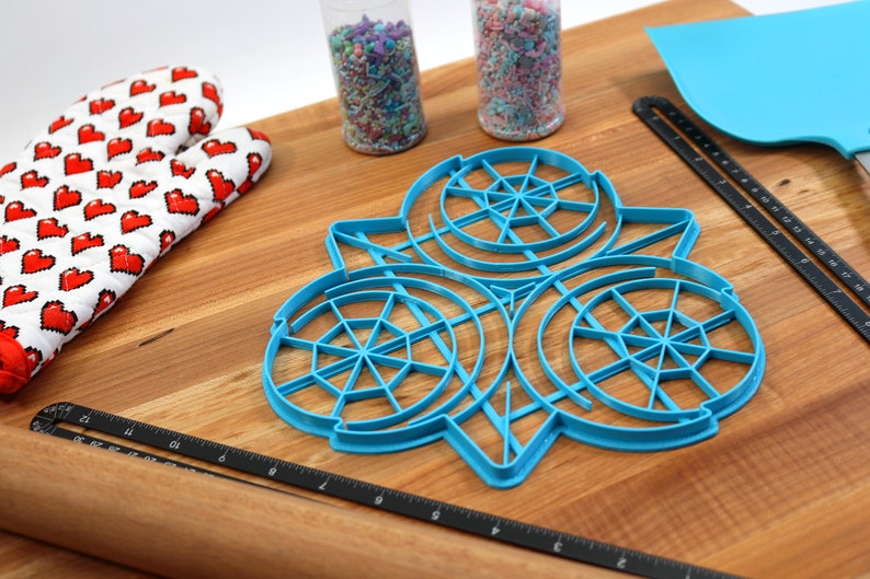 May include: A blue plastic cookie cutter shaped like three overlapping circles with a web-like design. The cutter is on a wooden surface with a rolling pin, a spatula, and two jars of sprinkles.