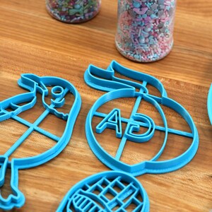 Puede incluir: Un juego de cortadores de galletas de pl&aacute;stico azul en forma de p&aacute;jaro, melocot&oacute;n con las letras "AD" en el interior, hoja y gofre. Los cortadores est&aacute;n sobre una superficie de madera.