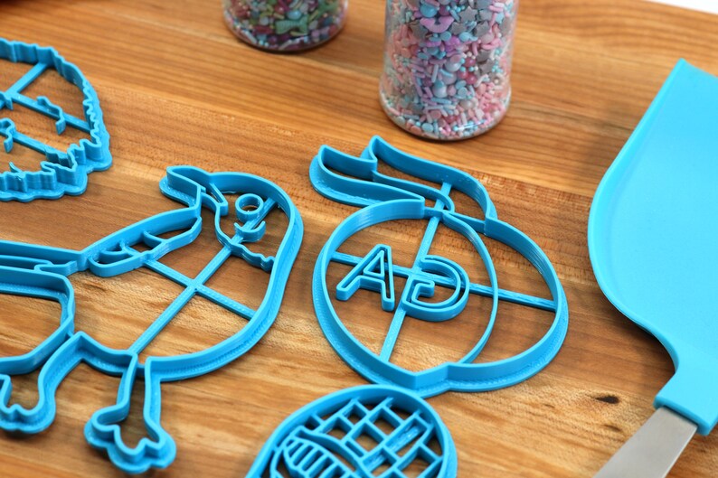 Puede incluir: Un juego de cortadores de galletas de pl&aacute;stico azul en forma de p&aacute;jaro, melocot&oacute;n con las letras "AD" en el interior, hoja y gofre. Los cortadores est&aacute;n sobre una superficie de madera.