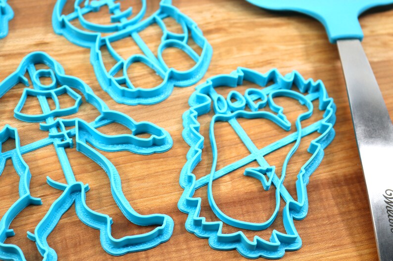 May include: A set of four blue plastic cookie cutters shaped like a bear, a footprint, a snowman, and a person. The cutters are on a wooden surface.