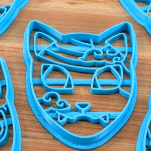 K&ouml;nnte beinhalten: Ein Satz mit vier blauen Plastik-Ausstechern in Katzenform. Die Ausstecher haben detaillierte Merkmale, darunter Augen, Nasen und Schnurrhaare.