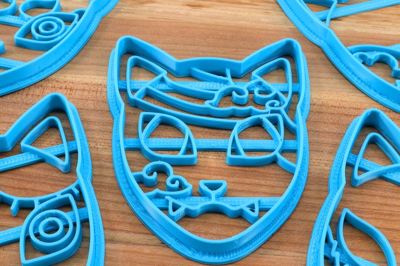 K&ouml;nnte beinhalten: Ein Satz mit vier blauen Plastik-Ausstechern in Katzenform. Die Ausstecher haben detaillierte Merkmale, darunter Augen, Nasen und Schnurrhaare.