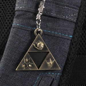May include: A gold-colored metal keychain featuring the Triforce symbol from the Legend of Zelda video game series. The Triforce is a triangular design with three points, each containing a different symbol. The keychain is attached to a silver chain.