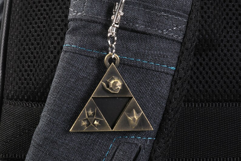 May include: A gold-colored metal keychain featuring the Triforce symbol from the Legend of Zelda video game series. The Triforce is a triangular design with three points, each containing a different symbol. The keychain is attached to a silver chain.