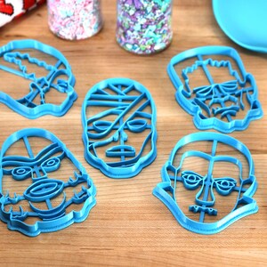 May include: A collection of five blue plastic cookie cutters in the shapes of monster faces, including a mummy, Frankenstein's monster, and a skull. The cutters are arranged on a wooden surface, with sprinkles and a spatula nearby.