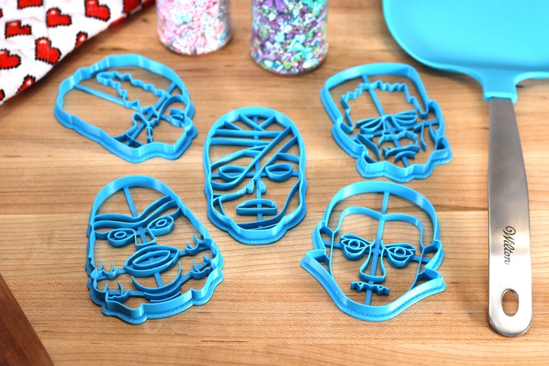 May include: A collection of five blue plastic cookie cutters in the shapes of monster faces, including a mummy, Frankenstein's monster, and a skull. The cutters are arranged on a wooden surface, with sprinkles and a spatula nearby.