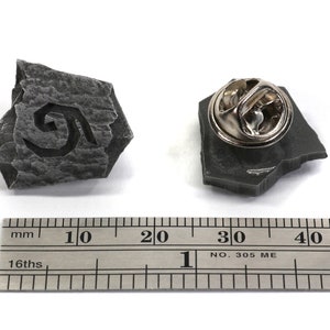 Puede incluir: Un pin de color gris en forma de piedra con un dise&ntilde;o en espiral. El pin mide aproximadamente 2,5 cm de di&aacute;metro y tiene un cierre met&aacute;lico en la parte posterior.