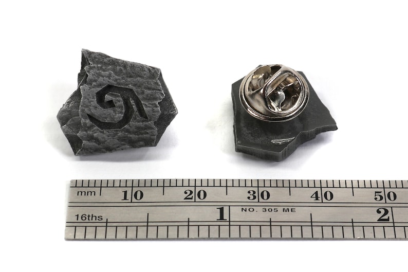 Puede incluir: Un pin de color gris en forma de piedra con un dise&ntilde;o en espiral. El pin mide aproximadamente 2,5 cm de di&aacute;metro y tiene un cierre met&aacute;lico en la parte posterior.