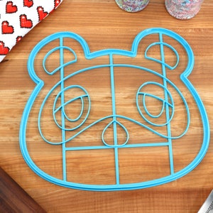 May include: Blue plastic cookie cutter shaped like a cartoon animal face. The cutter has a grid pattern inside the outline of the face.