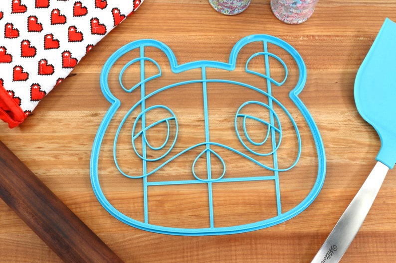 May include: Blue plastic cookie cutter shaped like a cartoon animal face. The cutter has a grid pattern inside the outline of the face.