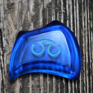 May include: A blue glass charm with a teal triquetra symbol. The charm is shaped like a crescent moon and has a smooth, glossy finish.