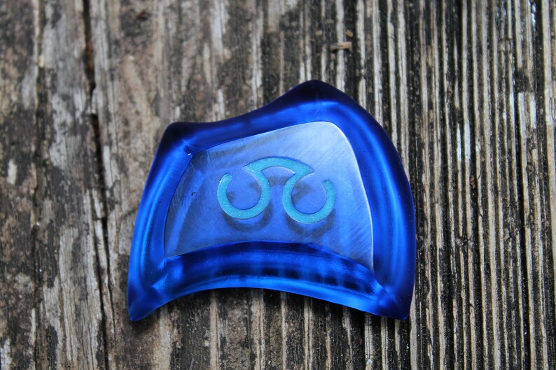 May include: A blue glass charm with a teal triquetra symbol. The charm is shaped like a crescent moon and has a smooth, glossy finish.