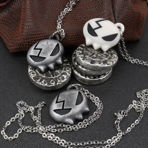 May include: Three silver necklaces with skull-shaped pendants. Two of the pendants are silver and one is white. The pendants have a black design on them. The necklaces are on a brown leather surface.