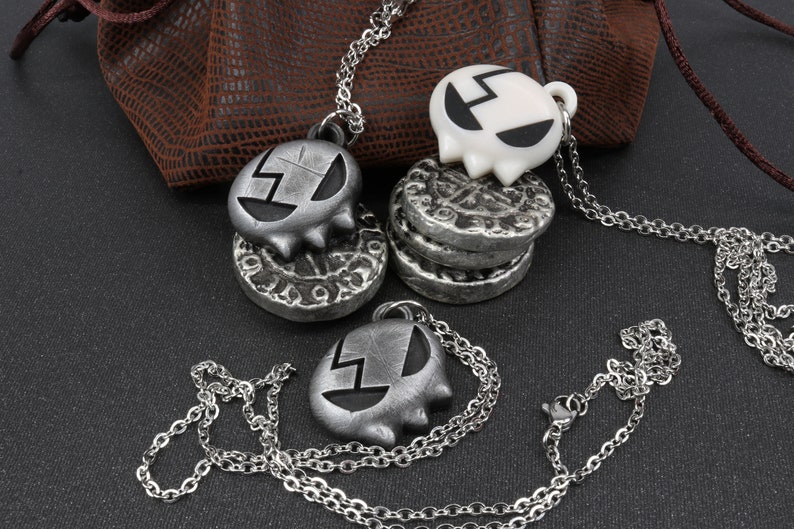 May include: Three silver necklaces with skull-shaped pendants. Two of the pendants are silver and one is white. The pendants have a black design on them. The necklaces are on a brown leather surface.
