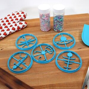 Op de afbeelding: Vijf blauwe koekjesvormen met verschillende ontwerpen, twee potten met kleurrijke sprinkles, een houten deegroller, een blauwe spatel en een rood-witte ovenwant op een houten oppervlak.