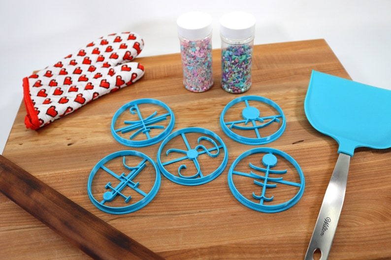 Op de afbeelding: Vijf blauwe koekjesvormen met verschillende ontwerpen, twee potten met kleurrijke sprinkles, een houten deegroller, een blauwe spatel en een rood-witte ovenwant op een houten oppervlak.