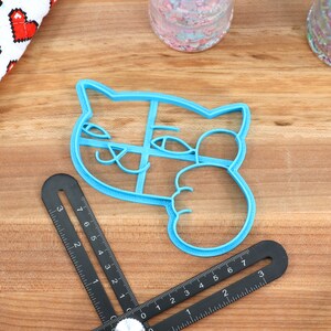 May include: A blue plastic cookie cutter shaped like a cat with its paw raised in a wave. The cutter has a detailed design with eyes, a nose, and whiskers.