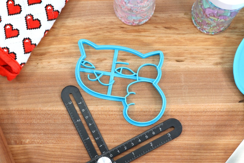May include: A blue plastic cookie cutter shaped like a cat with its paw raised in a wave. The cutter has a detailed design with eyes, a nose, and whiskers.
