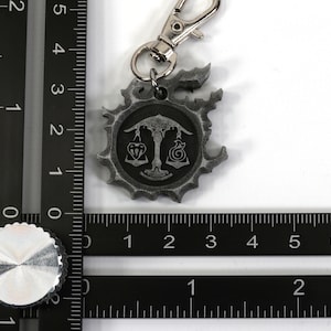 May include: A silver-colored keychain with a circular charm featuring a scale design and a symbol. The charm is attached to a silver-colored spring clasp.