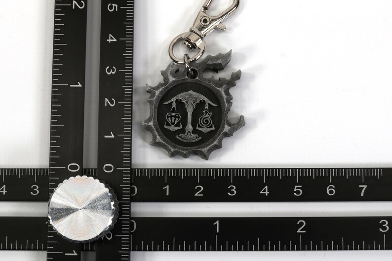 May include: A silver-colored keychain with a circular charm featuring a scale design and a symbol. The charm is attached to a silver-colored spring clasp.