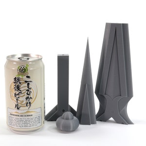 May include: A can of Japanese beer with the text "こしひかり" and "越後美人" on the label.  The can is next to three gray 3D printed objects: a cone, a rectangular prism, and a more complex object with a pointed top and a base that resembles a cross.