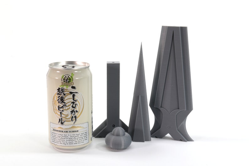 May include: A can of Japanese beer with the text "こしひかり" and "越後美人" on the label.  The can is next to three gray 3D printed objects: a cone, a rectangular prism, and a more complex object with a pointed top and a base that resembles a cross.
