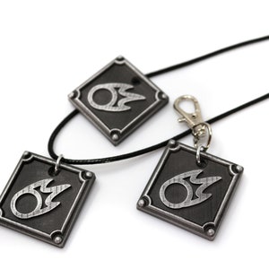 May include: Three silver-colored pendants with a black flame design on a black background. The pendants are attached to black cords, one of which is a necklace and the other two are keychains.