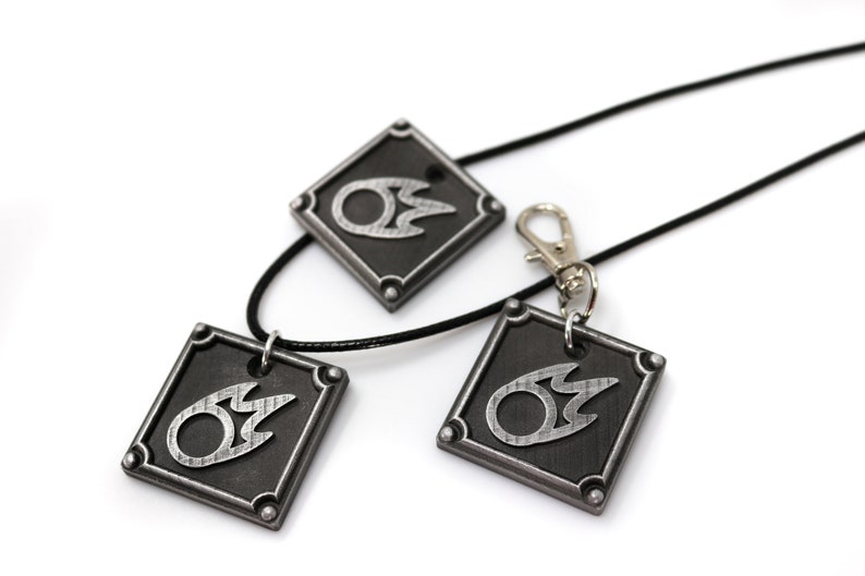 May include: Three silver-colored pendants with a black flame design on a black background. The pendants are attached to black cords, one of which is a necklace and the other two are keychains.