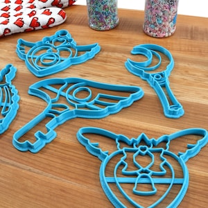 May include: Five blue plastic cookie cutters shaped like a crescent moon wand, a winged heart, a winged key, a winged heart with a crown, and a winged heart with a bow.