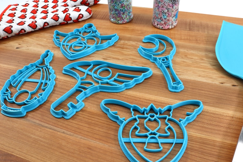 May include: Five blue plastic cookie cutters shaped like a crescent moon wand, a winged heart, a winged key, a winged heart with a crown, and a winged heart with a bow.