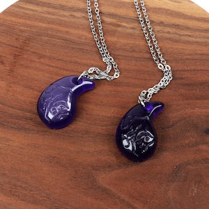 May include: Two silver chains with purple glass pendants shaped like a crescent moon. The pendants have intricate designs carved into them.
