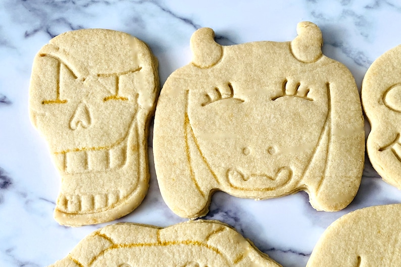 May include: Four sugar cookies decorated with icing. The cookies are shaped like a skull, a goat, a heart, and a flower. The cookies are on a white marble surface.