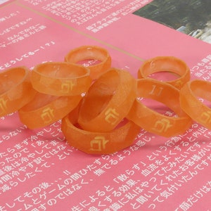 May include: A close-up of a group of orange plastic rings with a Japanese character printed on each ring. The rings are arranged in a pile on a pink surface.