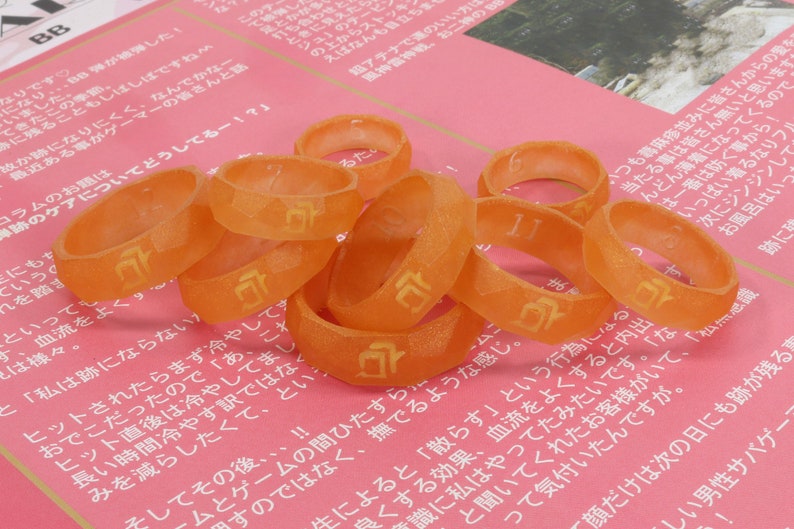 May include: A close-up of a group of orange plastic rings with a Japanese character printed on each ring. The rings are arranged in a pile on a pink surface.