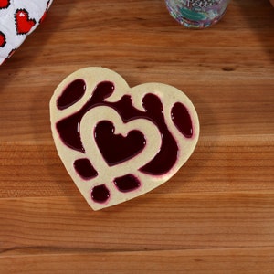 May include: A heart-shaped cookie decorated with a red jelly filling. The cookie is on a wooden surface.