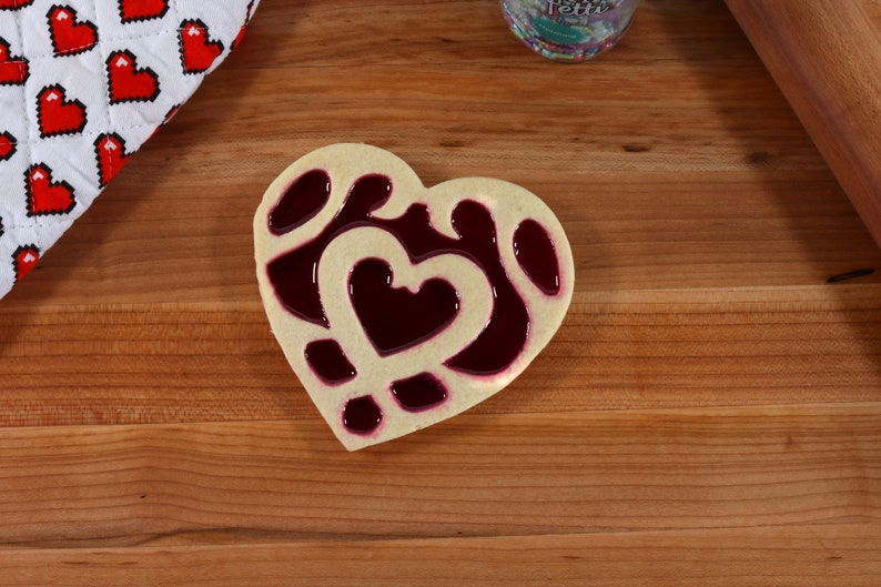 May include: A heart-shaped cookie decorated with a red jelly filling. The cookie is on a wooden surface.