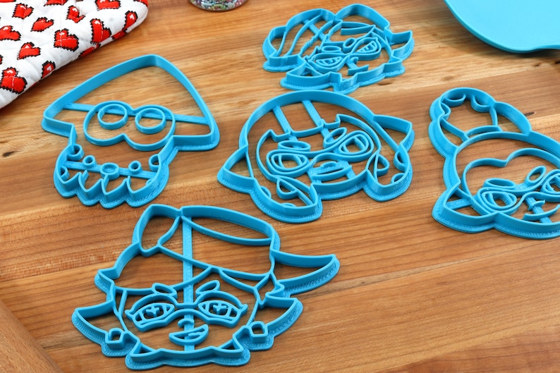May include: Five blue plastic cookie cutters shaped like cartoon characters. The characters have large eyes and are wearing hats.