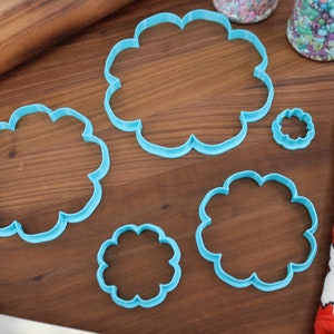 May include: A set of five blue plastic cookie cutters shaped like clouds. The cutters are arranged on a wooden surface.