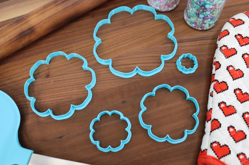 May include: A set of five blue plastic cookie cutters shaped like clouds. The cutters are arranged on a wooden surface.