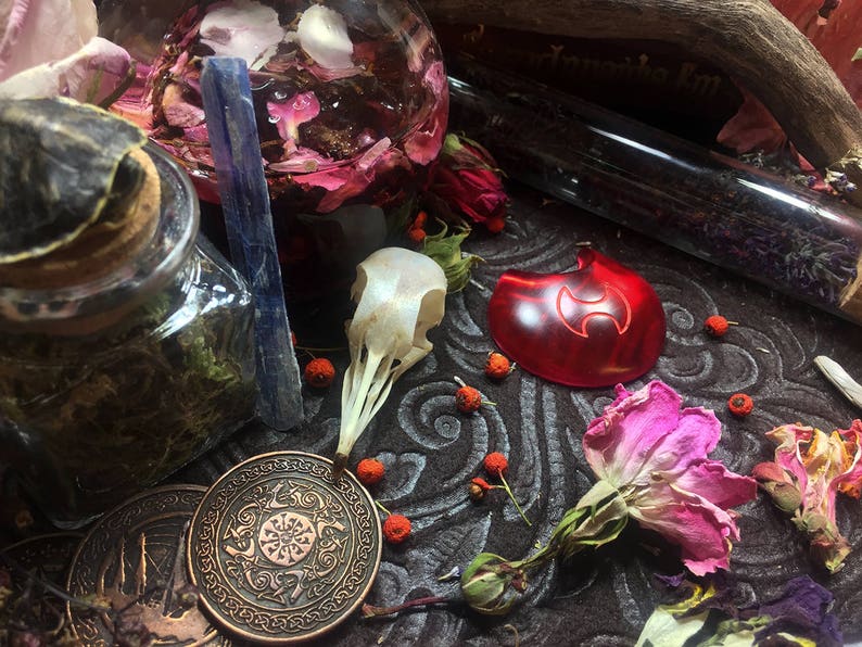 May include: A collection of witchy items including a glass jar filled with dried herbs, a glass bottle with dried flowers, a blue crystal, a red glass pendant with a symbol, a dried flower, and a white bone.