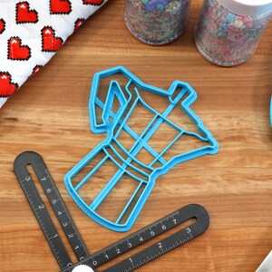 May include: Blue plastic cookie cutter shaped like a coffee pot. The cutter is on a wooden surface with other baking supplies.