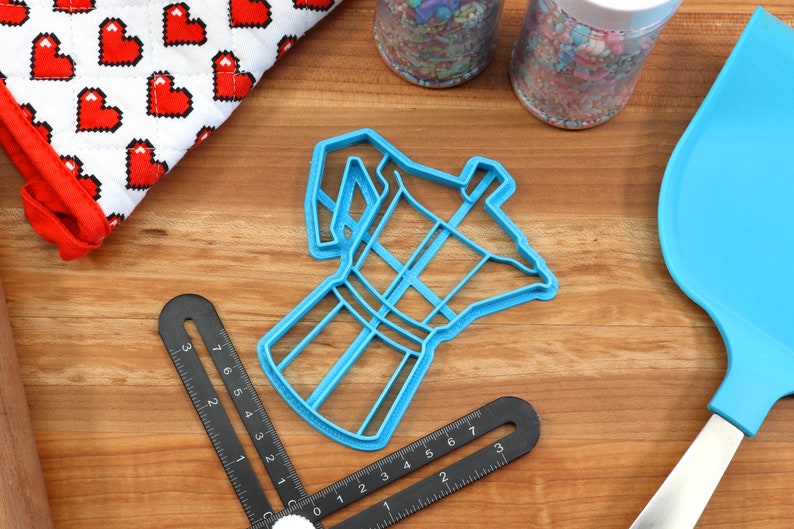 May include: Blue plastic cookie cutter shaped like a coffee pot. The cutter is on a wooden surface with other baking supplies.